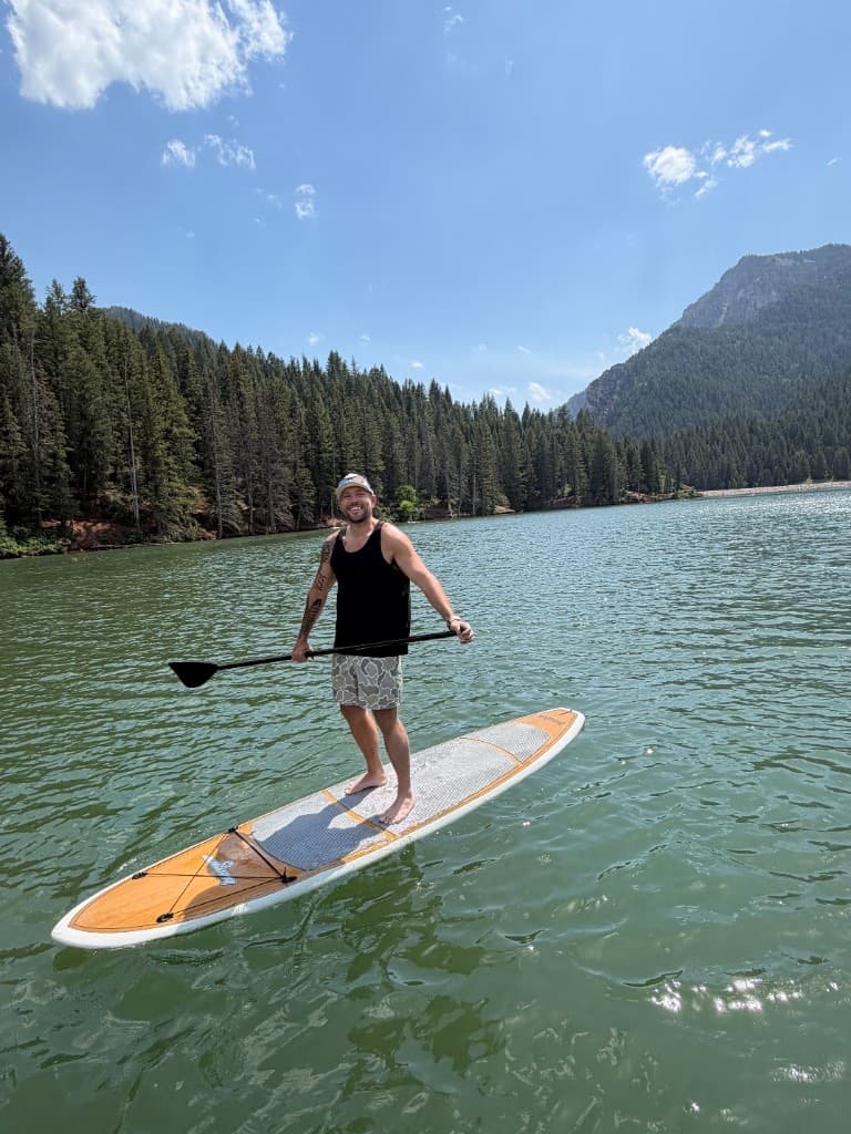 Nick standing on a paddleboard on an alpine lake ringed by pine forest and mountains