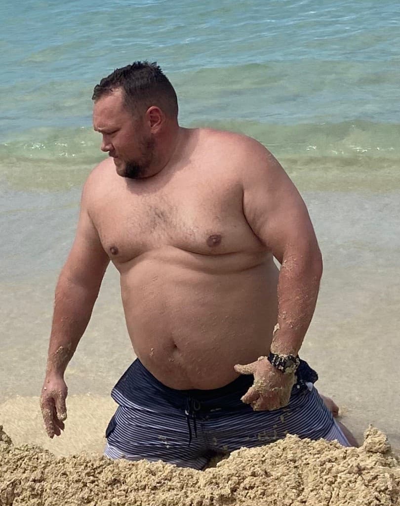 Nick shirtless on the beach at a heavier weight, kneeling in the sand